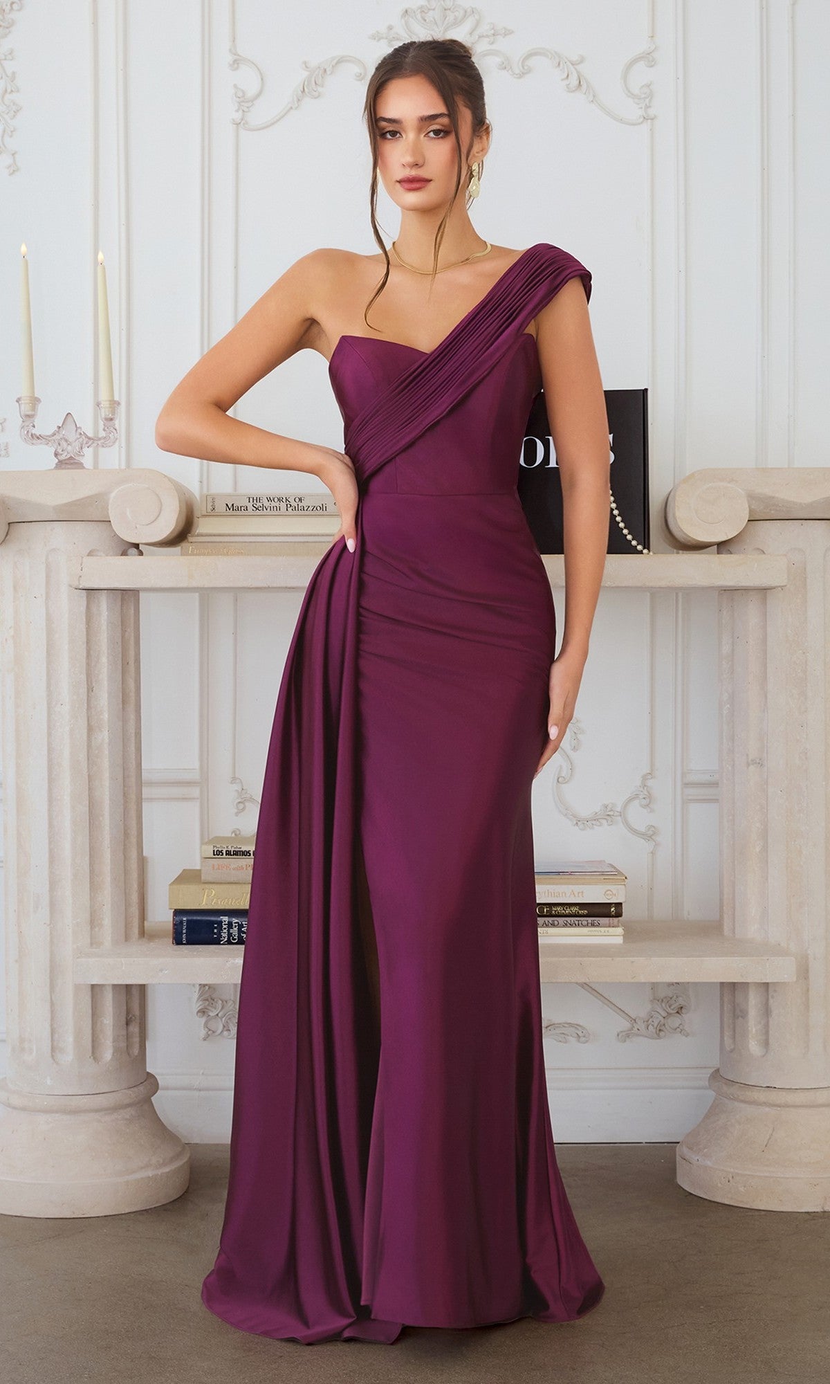 A woman stands indoors by a white mantel with books and candles, wearing the Amoris T817 One-Shoulder Long Chiffon Prom Dress in eggplant.